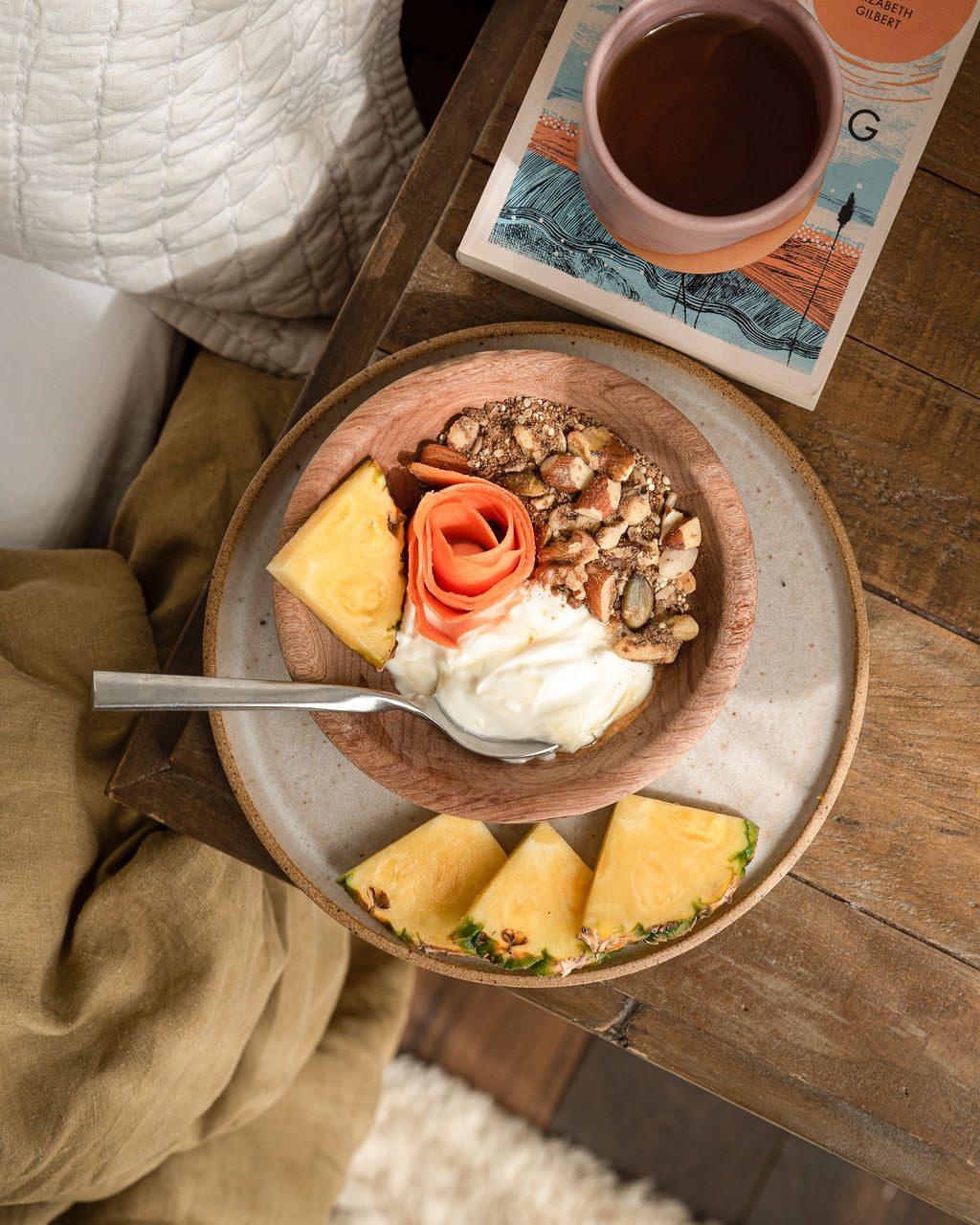 A bowl of Treasured Morning high protein granola on a table with yoghurt and fresh pineapple