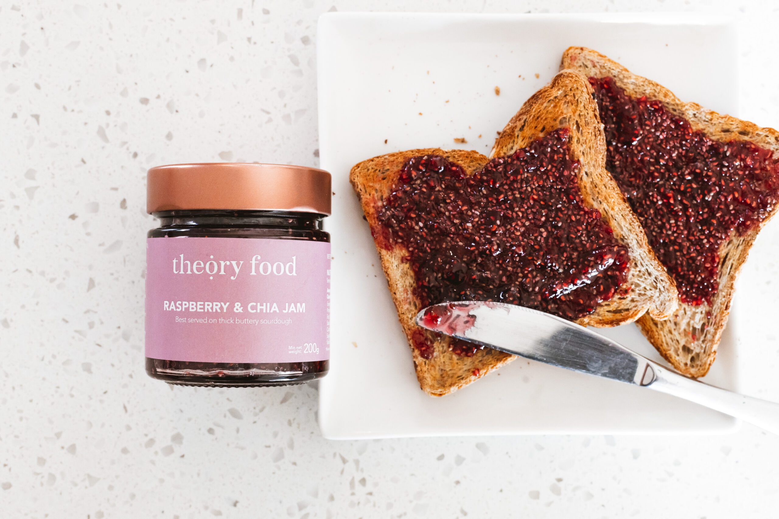Jar of Theory Food Raspberry and Chia Jam next to two slices of toast spread with the jam